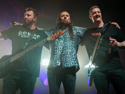 Three members of Thy Art is Murder are standing with their arms around each others shoulder whilst being back lit by purple and white lights. On the left is a bassist, in the middle is the vocalist, and on the right is the guitarist. All three members are part of an Australian death metal band.